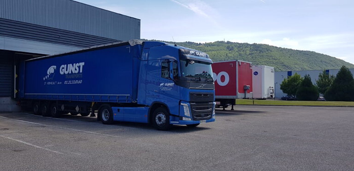Un camion semi-remorque bleu à bâche coulissante, marqué Gunst, est garé sur un parking devant un entrepôt, avec une colline boisée à l'arrière-plan.