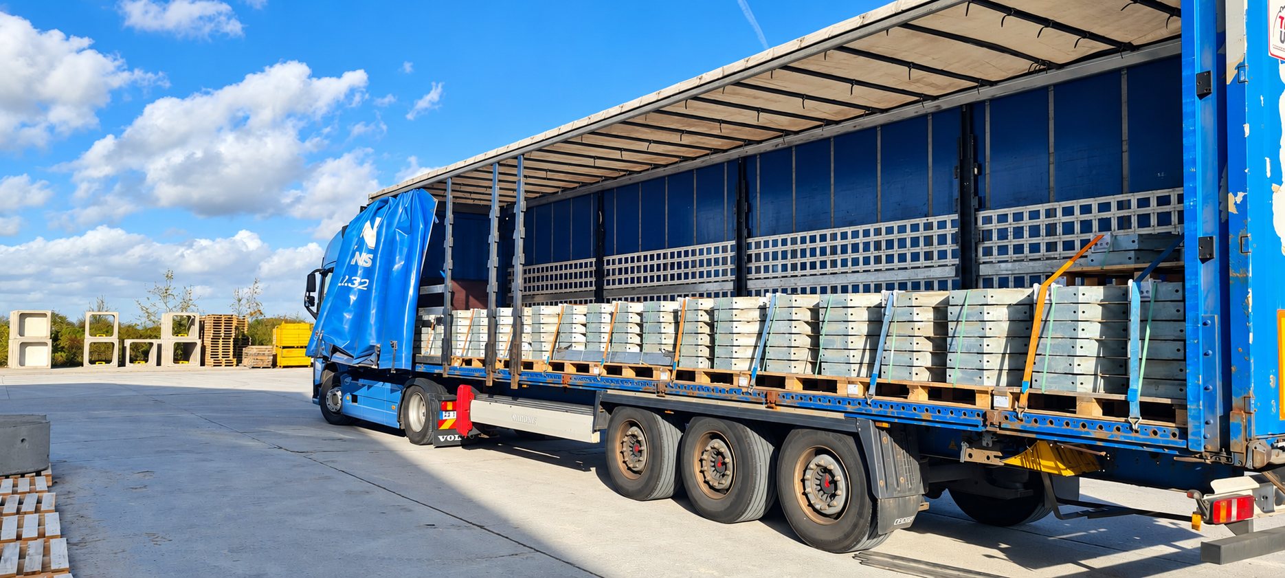 Un camion bleu aux rideaux coulissants ouverts, chargé de palettes de dalles en béton, stationné sur une aire de stockage ensoleillée.