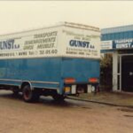 Un camion de déménagement bleu et blanc portant le nom Gunst est garé sur un parking devant un bâtiment commercial.
