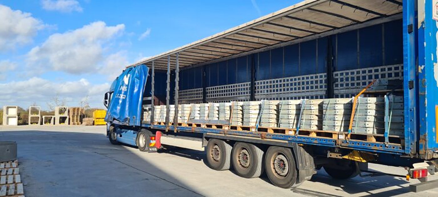 Un camion bleu à plateau bâché est chargé de dalles en béton empilées sur des palettes, sur une aire de stockage ensoleillée.