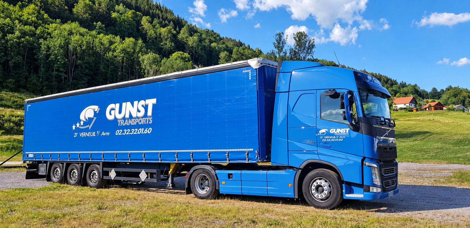 Un camion Volvo bleu Gunst avec sa remorque à bâches, garé sur l’herbe devant une colline boisée, sous un ciel bleu.