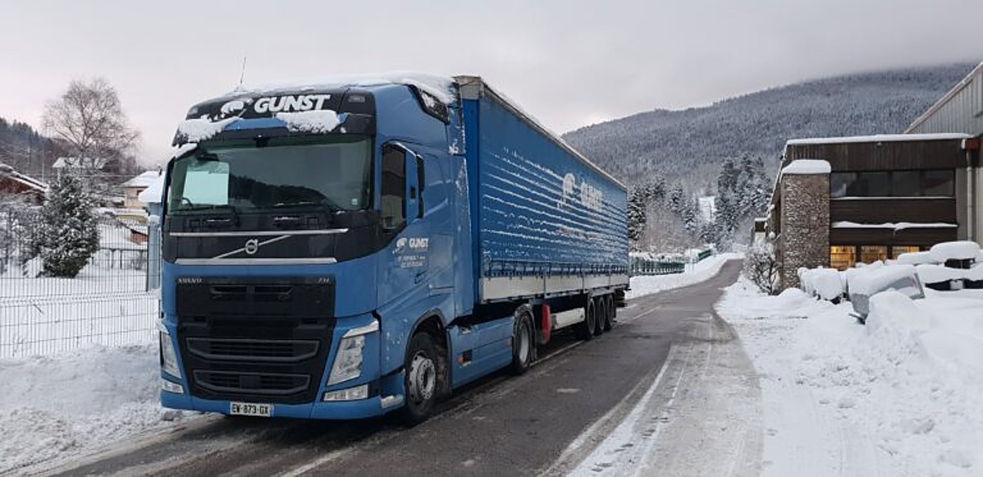 Un camion Volvo bleu aux couleurs de Gunst, avec sa remorque à bâches, est garé sur une route de montagne couverte de neige.