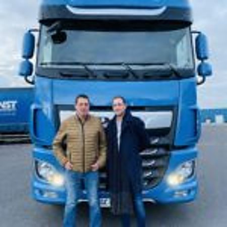 Deux hommes se tiennent côte à côte devant la cabine d'un camion bleu garé à l'extérieur, sous un ciel dégagé.